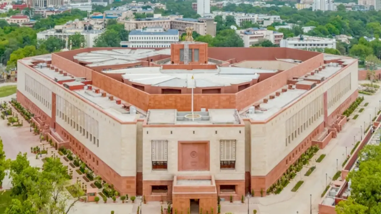 Aerial view of India's new Parliament House in New Delhi, featuring its triangular design, red sandstone exterior, and the national emblem atop the central structure.
